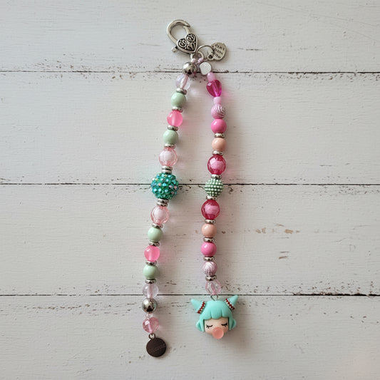 A handmade beaded keychain or phone strap with a silver lobster clasp, featuring pink, mint green, and metallic beads, a green beaded ball accent, a small black charm, and a mint green anime-style character charm at the bottom, displayed against a white wood background.