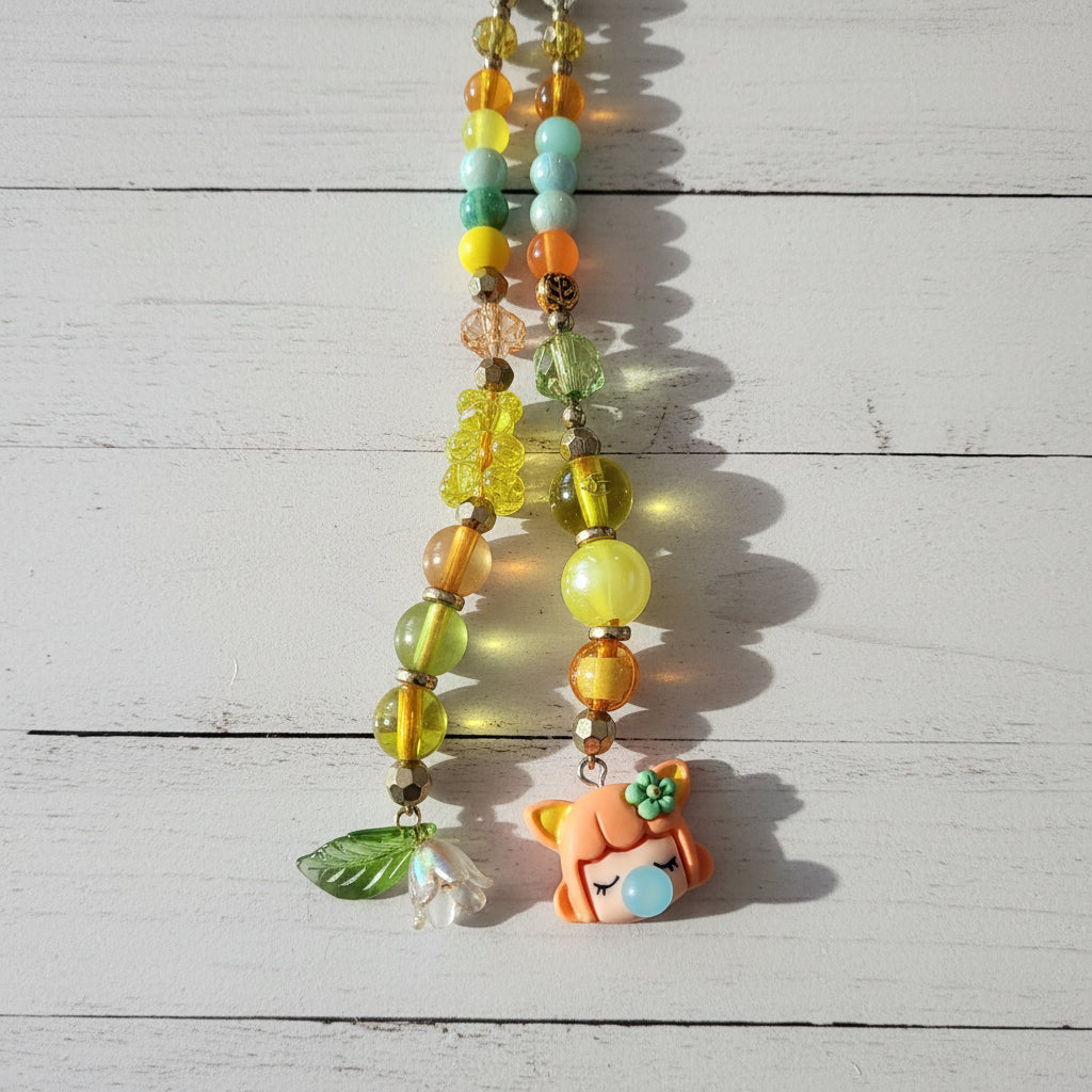 A close-up of a beaded necklace or phone strap laid on a light wood surface. The piece features translucent yellow and green beads, metallic gold-toned beads, a small green leaf charm, and a cute orange cartoon character charm with a blue bubblegum bubble.