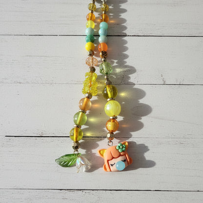 A close-up of a beaded necklace or phone strap laid on a light wood surface. The piece features translucent yellow and green beads, metallic gold-toned beads, a small green leaf charm, and a cute orange cartoon character charm with a blue bubblegum bubble.