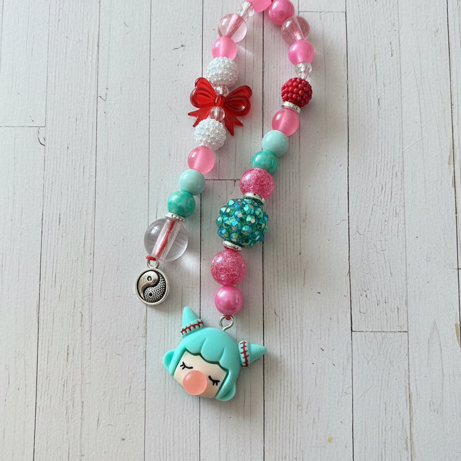 A beaded necklace featuring a green mermaid character charm with closed eyes, pink and green beads, red and white accents, and a silver crescent moon charm, displayed on a light wood plank background.
