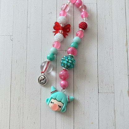 A beaded necklace featuring a green mermaid character charm with closed eyes, pink and green beads, red and white accents, and a silver crescent moon charm, displayed on a light wood plank background.
