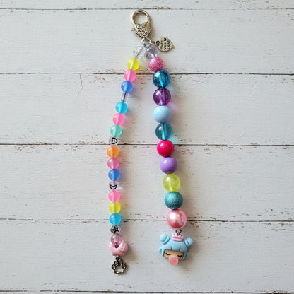A colorful beaded phone strap or keychain charm hanging against a light wood plank background. The strap features two strands: one with translucent neon pink, blue, yellow, and orange beads, and one with glossy blue, purple, pink, and light blue beads, both ending in pastel charms including a pink bunny and a light blue creature.
