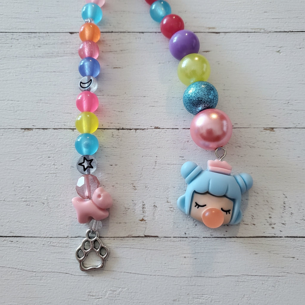 A beaded necklace or charm strand laid flat on a light wood surface, featuring colorful translucent beads, a pink charm, a silver paw print charm, and a blue character charm with an orange nose.