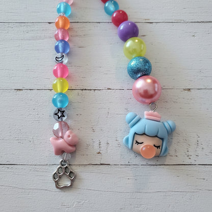 A beaded necklace or charm strand laid flat on a light wood surface, featuring colorful translucent beads, a pink charm, a silver paw print charm, and a blue character charm with an orange nose.