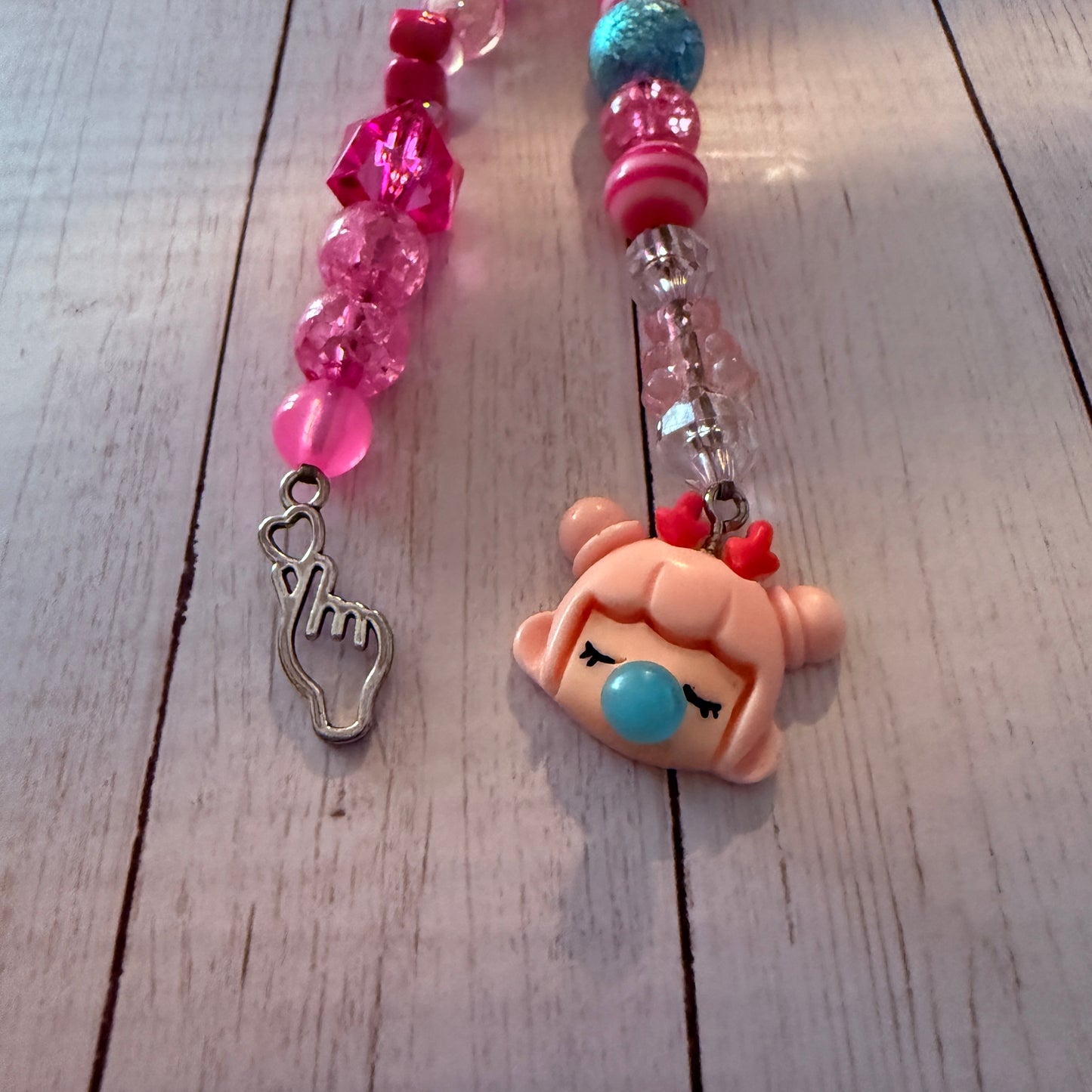 A close-up of a beaded charm or keychain laid on a light wood surface. The left side has pink faceted beads and a silver heart-hand gesture charm.