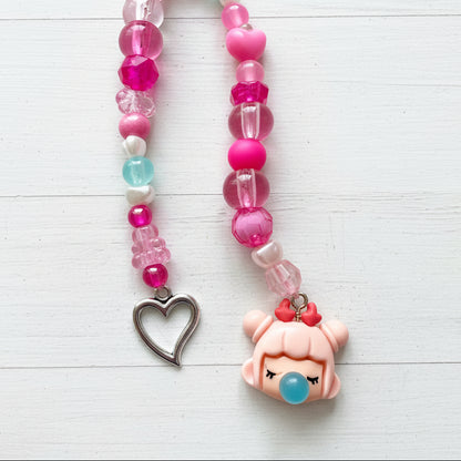 A beaded necklace or phone strap laid on a light wood surface, featuring pink and clear faceted beads, a silver heart-shaped charm, and a pink cartoon character charm with a blue nose and red bow.