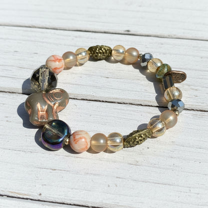 A beaded stretch bracelet featuring a mix of metallic gold, iridescent blue, peach, and gray glass beads, displayed on a white wooden surface.