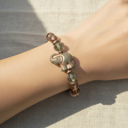 A beaded bracelet featuring a small copper elephant charm, faceted beads in brown, green, and cream, and metallic spacer beads, displayed on a lady's arm