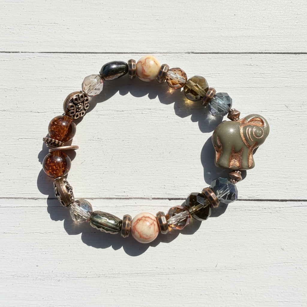 A beaded stretch bracelet featuring a carved elephant bead, faceted glass beads in amber, brown, and blue-gray, and a copper-toned filigree accent, displayed on a weathered white wooden surface.