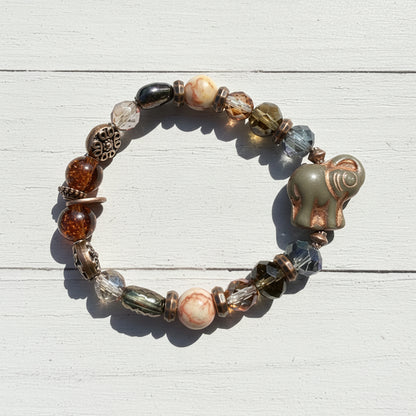 A beaded stretch bracelet featuring a carved elephant bead, faceted glass beads in amber, brown, and blue-gray, and a copper-toned filigree accent, displayed on a weathered white wooden surface.