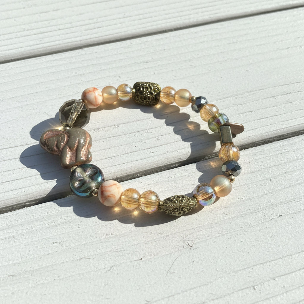 A beaded stretch bracelet featuring a mix of translucent glass beads, metallic gold-toned accents, and a carved elephant bead, displayed on a weathered white wood surface.