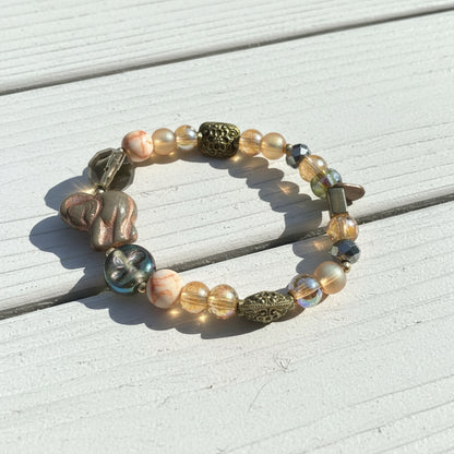 A beaded stretch bracelet featuring a mix of translucent glass beads, metallic gold-toned accents, and a carved elephant bead, displayed on a weathered white wood surface.