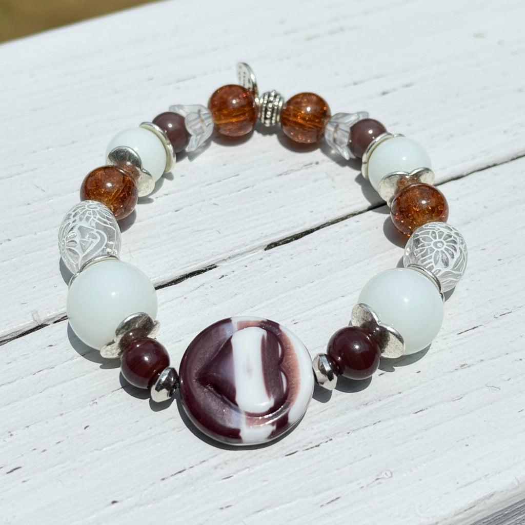 A beaded stretch bracelet featuring white glass beads, amber brown translucent beads, silver filigree beads, and a central marbled round bead with white and dark burgundy sections, displayed on a wooden surface.