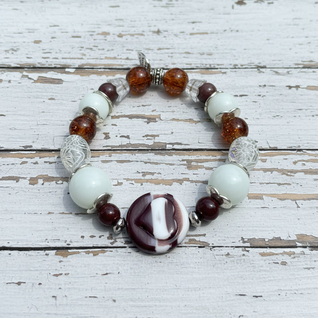 A beaded stretch bracelet featuring white glass beads, amber brown translucent beads, silver filigree beads, and a central marbled round bead with white and dark burgundy sections, displayed on a white wooden surface.