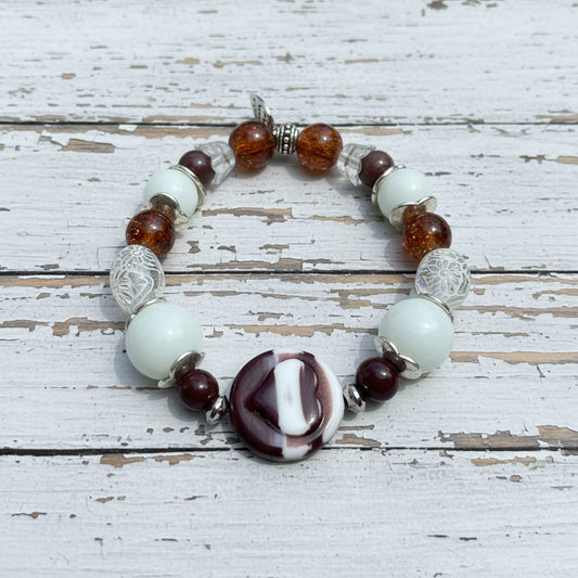 A beaded stretch bracelet featuring white glass beads, amber brown translucent beads, silver filigree beads, and a central marbled round bead with white and dark burgundy sections, displayed on a white wooden surface.