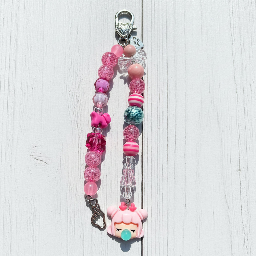 A pink beaded keychain or phone strap charm with a cartoon character charm, displayed on a white wooden surface.