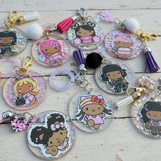 A collection of round acrylic keychains featuring cartoon characters, including Hello Kitty and Black Cat designs, decorated with glitter, sequins, tassel charms, and pom-poms on a tan background.