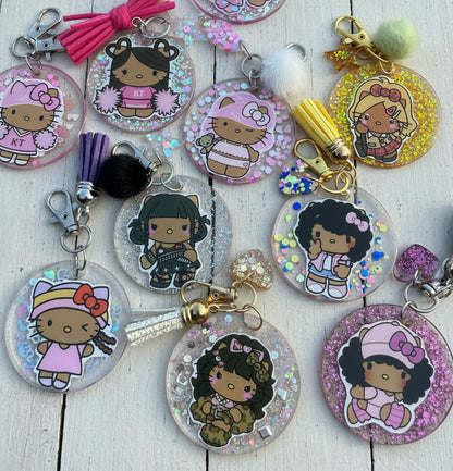 A group of round acrylic keychains featuring cartoon girl illustrations, some with glitter backgrounds and tassel charms, arranged on a white surface.