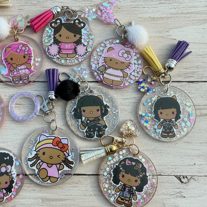 A close-up flat lay of multiple round acrylic keychains featuring cartoon Hello Kitty-style characters in pink outfits, decorated with glitter, tassels, pom-poms, and key rings on a white surface.