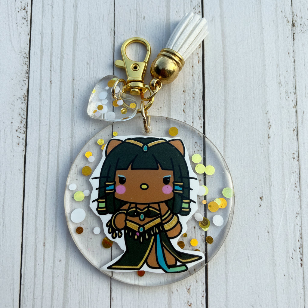 A round acrylic keychain charm featuring a cartoon character with black hair and a gold outfit, suspended in clear resin with gold and white glitter. The keychain includes a gold clasp, a white tassel, and a small clear heart charm on a brown kraft paper background.
