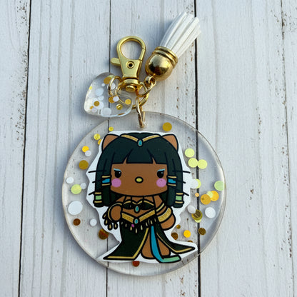 A round acrylic keychain charm featuring a cartoon character with black hair and a gold outfit, suspended in clear resin with gold and white glitter. The keychain includes a gold clasp, a white tassel, and a small clear heart charm on a brown kraft paper background.