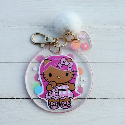A Hello Kitty acrylic keychain featuring a round clear charm with a pink and brown Hello Kitty character illustration, a white fluffy pom-pom, a silver clasp, and small pastel heart and gem accents, displayed on a white background.