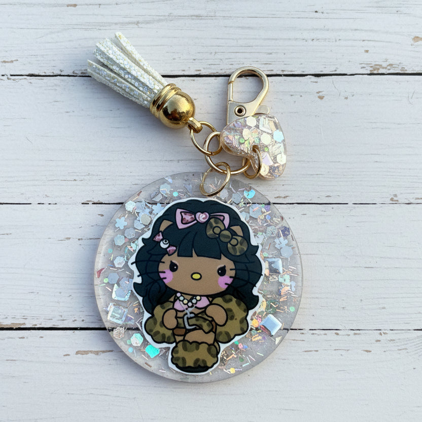 A round acrylic keychain charm featuring a cartoon character with black hair, a pink bow, and a leopard-print outfit, suspended in clear resin with iridescent glitter. The keychain includes a white tassel, gold-toned hardware, and a small heart-shaped charm, photographed on a white wooden background.