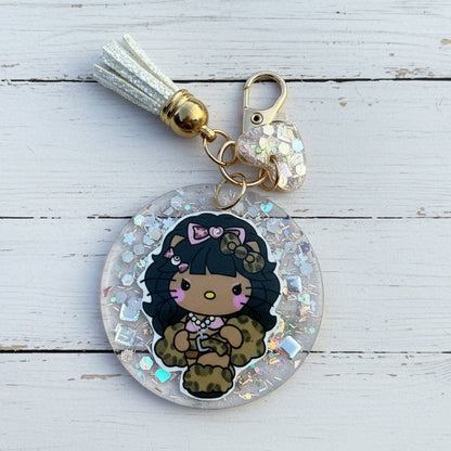 A round acrylic keychain charm featuring a cartoon character with black hair, a pink bow, and a leopard-print outfit, suspended in clear resin with iridescent glitter. The keychain includes a white tassel, gold-toned hardware, and a small heart-shaped charm, photographed on a white wooden background.