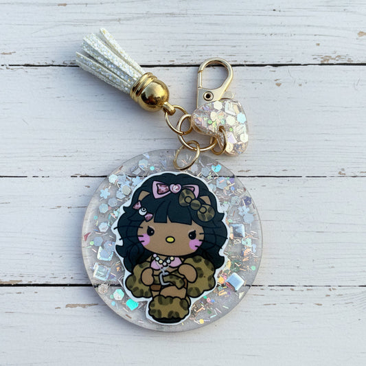 A round acrylic keychain charm featuring a cartoon character with black hair, a pink bow, and a leopard-print outfit, suspended in clear resin with iridescent glitter. The keychain includes a white tassel, gold-toned hardware, and a small heart-shaped charm, photographed on a white wooden background.