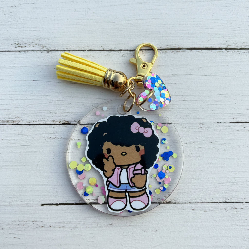 A round acrylic keychain featuring a cartoon girl with black hair and a pink bow, surrounded by colorful confetti. The keychain includes a yellow tassel, gold clasp, and a small heart-shaped charm with blue and pink confetti.