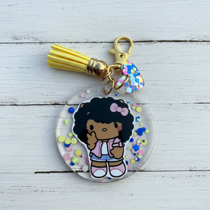 A round acrylic keychain featuring a cartoon girl with black hair and a pink bow, surrounded by colorful confetti. The keychain includes a yellow tassel, gold clasp, and a small heart-shaped charm with blue and pink confetti.