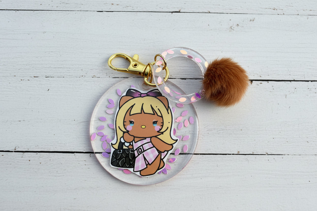 A round acrylic keychain charm featuring a cute cartoon girl character with blonde hair, a brown bow, and a pink-and-white dress, attached to a clear ring with iridescent dots, a gold clasp, and a brown faux-fur pom-pom, photographed on a tan textured background.