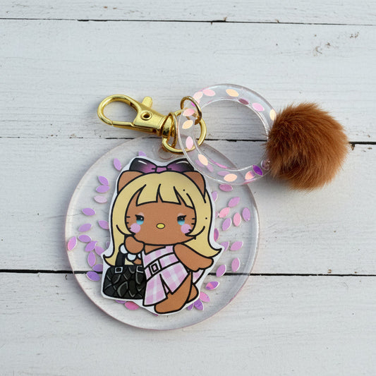A round acrylic keychain charm featuring a cute cartoon girl character with blonde hair, a brown bow, and a pink-and-white dress, attached to a clear ring with iridescent dots, a gold clasp, and a brown faux-fur pom-pom, photographed on a tan textured background.