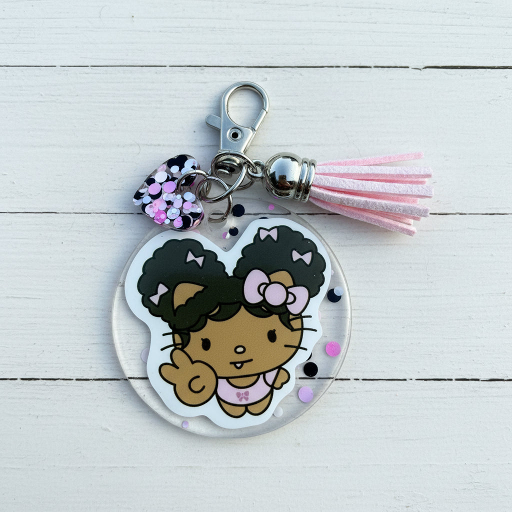 A Hello Kitty acrylic keychain featuring a cartoon character with black hair styled in two buns, pink bows, and a pink outfit, attached to a silver clasp with a pink tassel and a black-and-pink sequin charm, shown on a brown background.