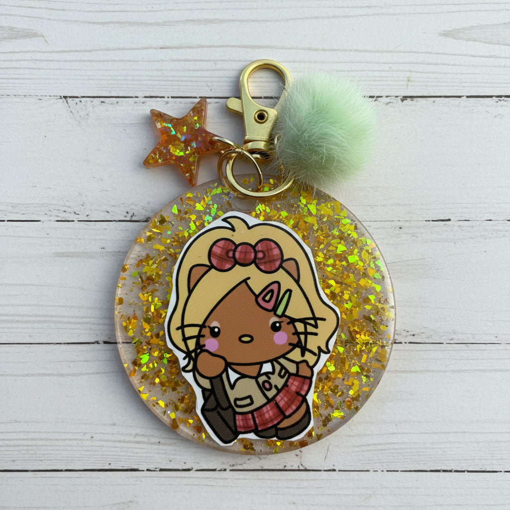 A round clear acrylic keychain charm filled with gold glitter, featuring a cartoon cat character sticker, a gold clasp, a translucent star, and a mint green pom-pom on a white background.