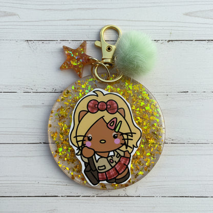 A round clear acrylic keychain charm filled with gold glitter, featuring a cartoon cat character sticker, a gold clasp, a translucent star, and a mint green pom-pom on a white background.