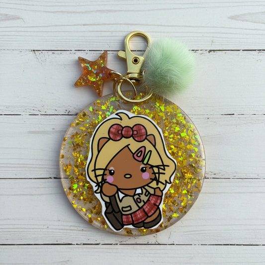 A round clear acrylic keychain charm filled with gold glitter, featuring a cartoon cat character sticker, a gold clasp, a translucent star, and a mint green pom-pom on a white background.