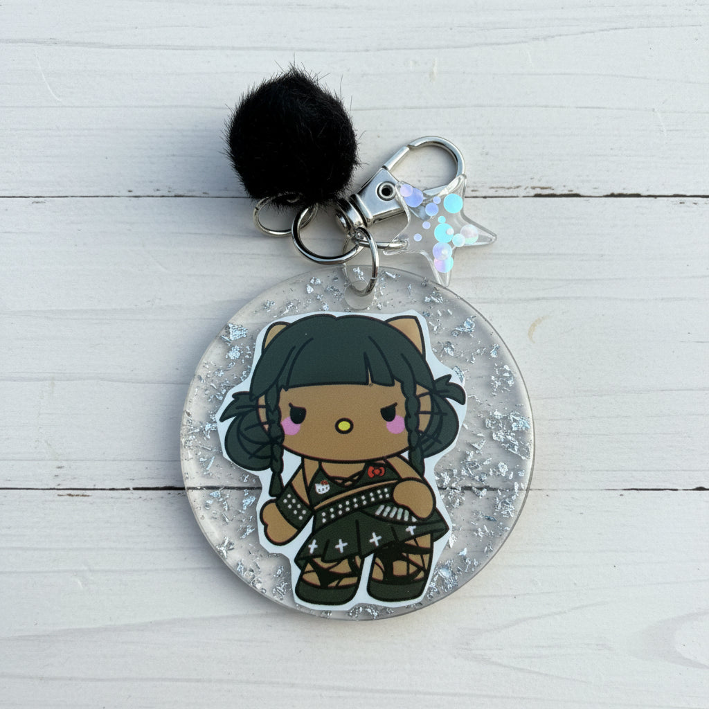 A round acrylic keychain featuring a chibi-style character with black hair and a black outfit, suspended inside clear glitter-filled resin. The keychain includes a black pom-pom, silver metal rings, and a small iridescent star charm, photographed on a brown kraft paper background.