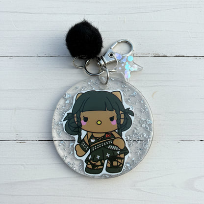 A round acrylic keychain featuring a chibi-style character with black hair and a black outfit, suspended inside clear glitter-filled resin. The keychain includes a black pom-pom, silver metal rings, and a small iridescent star charm, photographed on a brown kraft paper background.