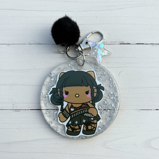 A round acrylic keychain featuring a chibi-style character with black hair and a black outfit, suspended inside clear glitter-filled resin. The keychain includes a black pom-pom, silver metal rings, and a small iridescent star charm, photographed on a brown kraft paper background.