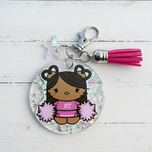 A personalized acrylic keychain charm featuring a cartoon cheerleader with the initials 'KT' on her pink shirt. The charm is circular with silver glitter and is attached to a silver lobster clasp, a silver star charm, and a bright pink tassel, all displayed on a white wooden background.
