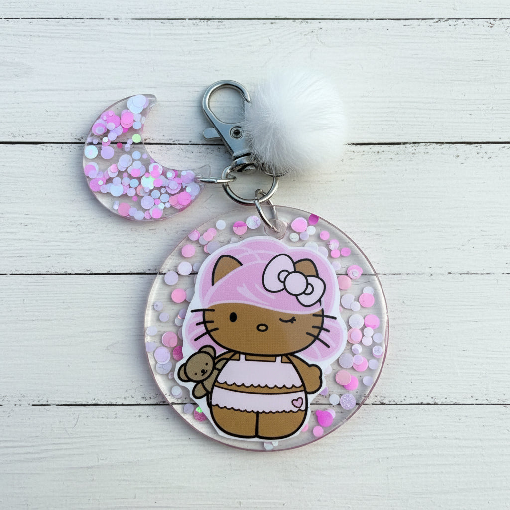 A round and crescent moon-shaped resin keychain charm featuring a Hello Kitty illustration with a pink and white striped dress, decorated with pink, white, and blue glitter, attached to a silver clasp with a white pom-pom on a white wooden background.