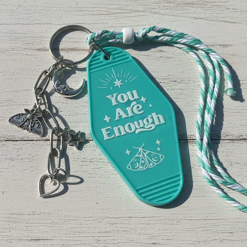 A turquoise plastic keychain with the words 'You Are Enough' and moon, star, and butterfly charms, attached to a mint and white twisted cord, photographed on a brown kraft paper background.