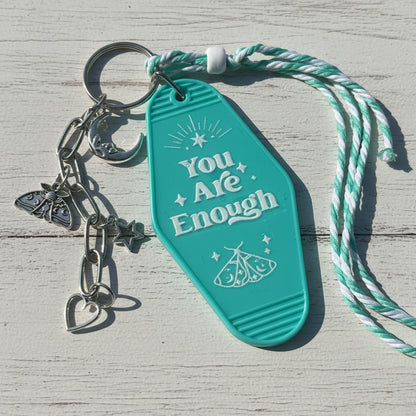 A turquoise plastic keychain with the words 'You Are Enough' and moon, star, and butterfly charms, attached to a mint and white twisted cord, photographed on a brown kraft paper background.