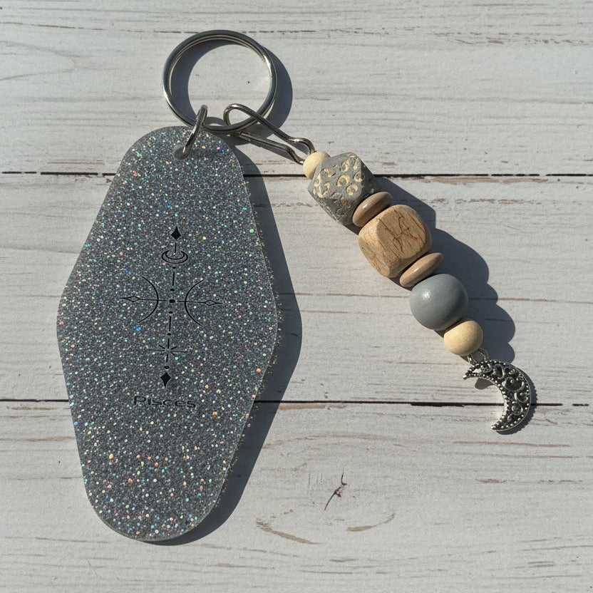 A keychain featuring a large, hexagonal silver glitter pendant engraved with a compass and celestial symbols, attached to a keyring with wooden and stone beads and a silver crescent moon charm, set against a white wooden background.