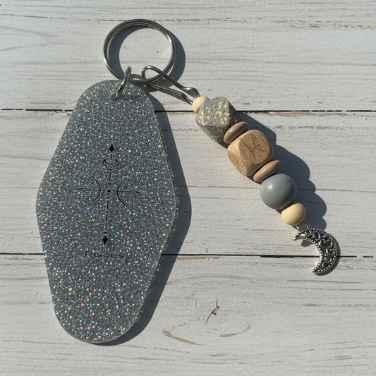 A keychain featuring a large, hexagonal silver glitter pendant engraved with a compass and celestial symbols, attached to a keyring with wooden and stone beads and a silver crescent moon charm, set against a white wooden background.