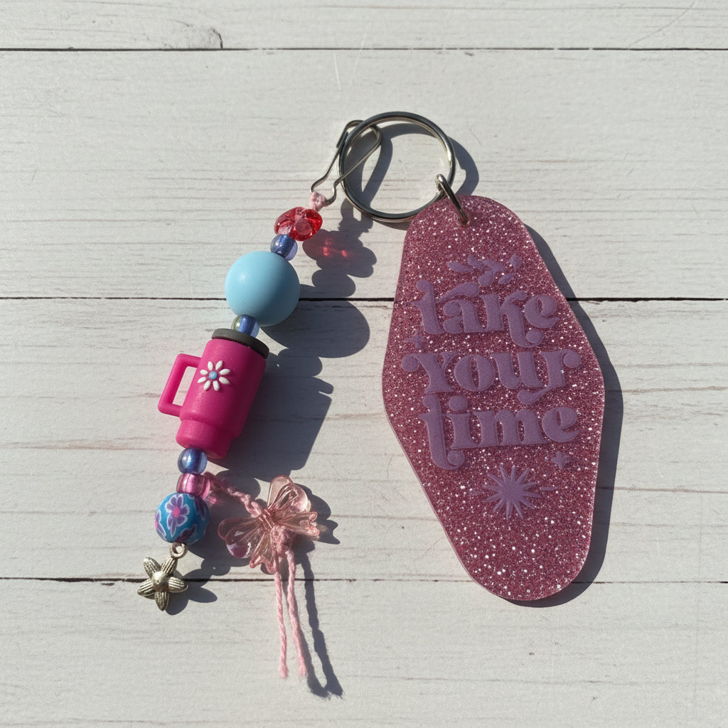 A pink glitter keychain with the phrase 'take your time' embossed on the main tag, shown alongside decorative beads, charms, and a metal key ring on a white background.
