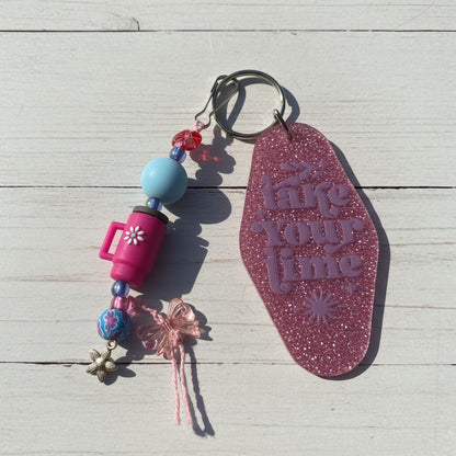 A pink glitter keychain with the phrase 'take your time' embossed on the main tag, shown alongside decorative beads, charms, and a metal key ring on a white background.