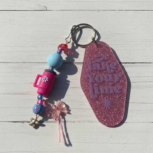 A pink glitter keychain with the phrase 'take your time' embossed on the main tag, shown alongside decorative beads, charms, and a metal key ring on a white background.