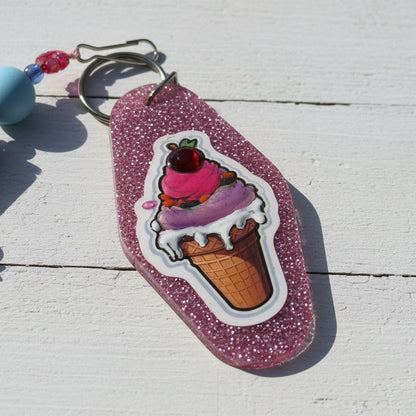 A pink glitter keychain featuring a die-cut sticker of an ice cream cone with pink and purple ice cream, white dripping sauce, and a cherry on top, shown on a white wood background.